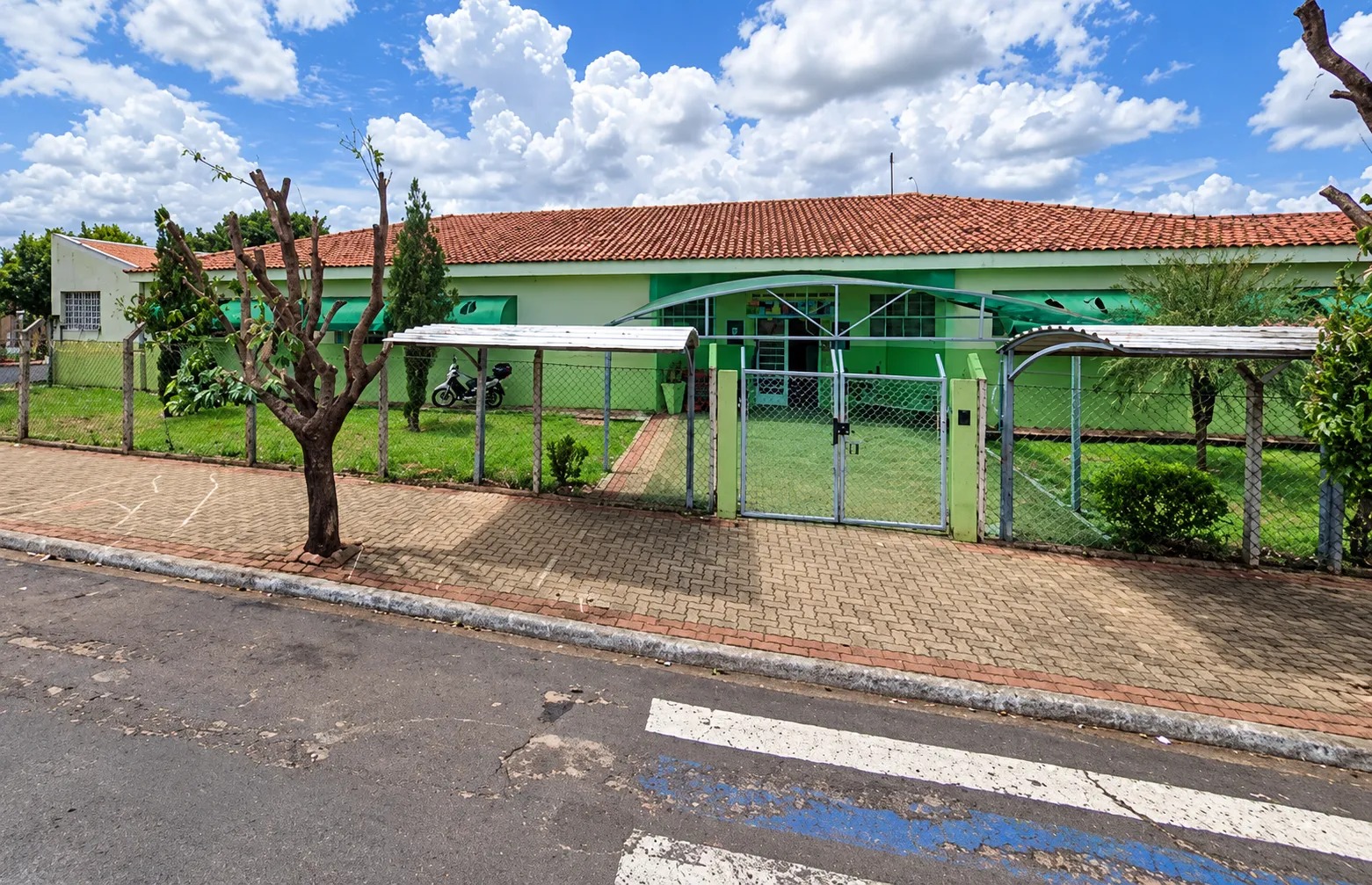 Prédio térreo de escola pintado de verde, com telhado de telha e portão de grade, visto da rua