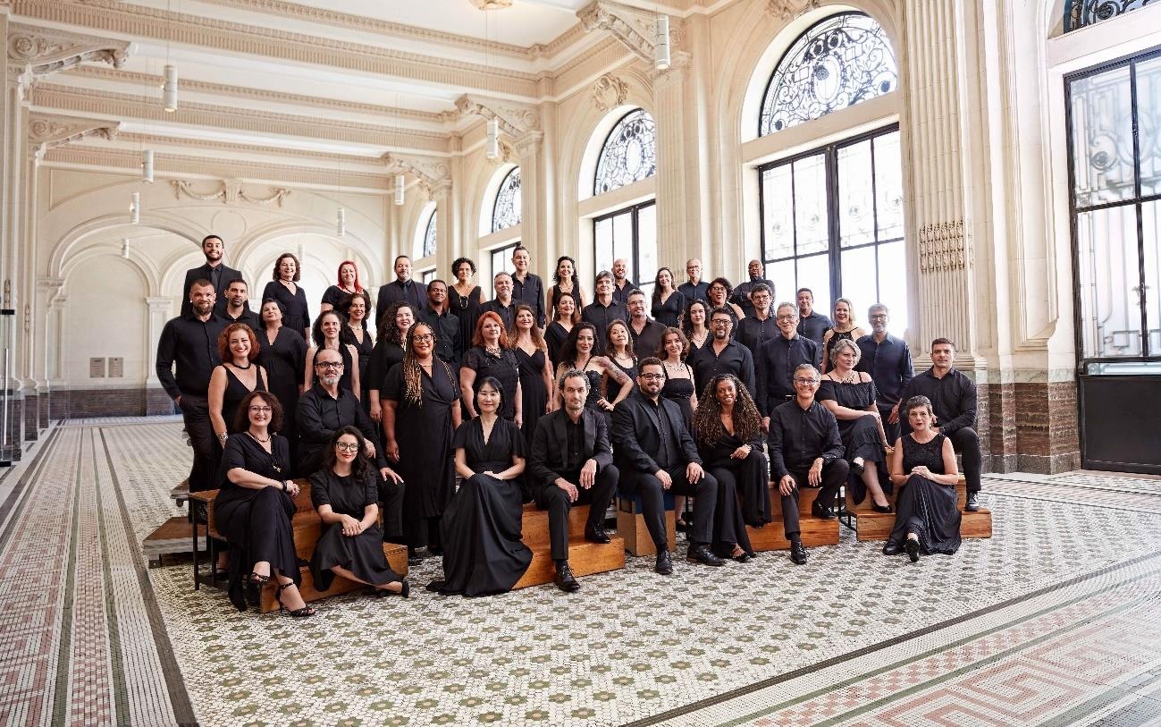 Integrantes do Coro da Osesp, vestidos de preto, posam sentados e em pé em um salão com janelas em arco