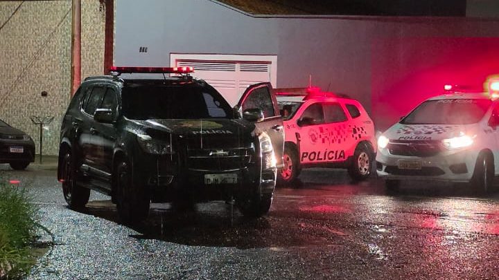 Homem de camiseta e calça escura caído no asfalto de uma rua, com carro preto estacionado ao lado e viaturas da Polícia Militar.