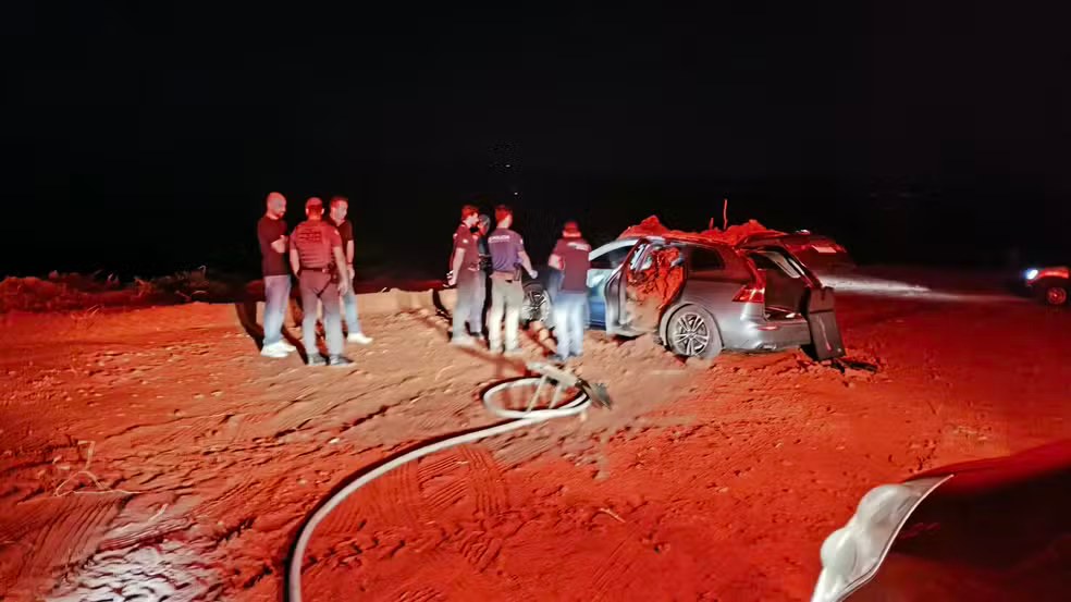 Policiais ao lado de um SUV coberto de terra, à noite, em estrada de terra