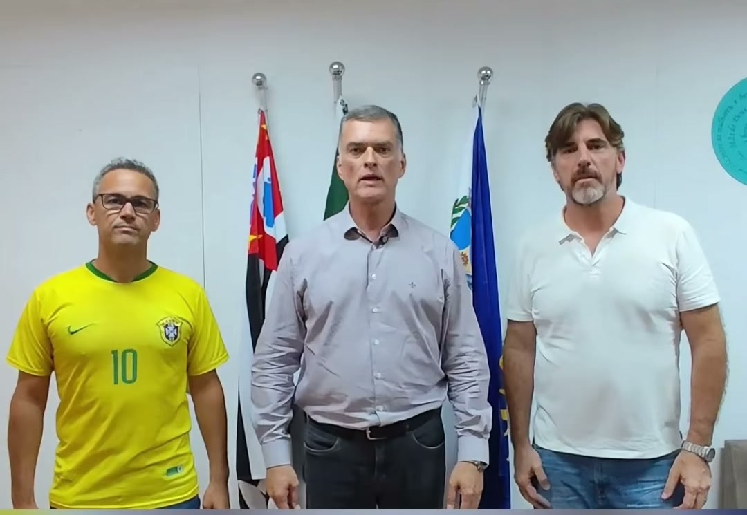 Três homens em pé lado a lado diante de bandeiras; o do centro, de camisa social, fala para a câmera