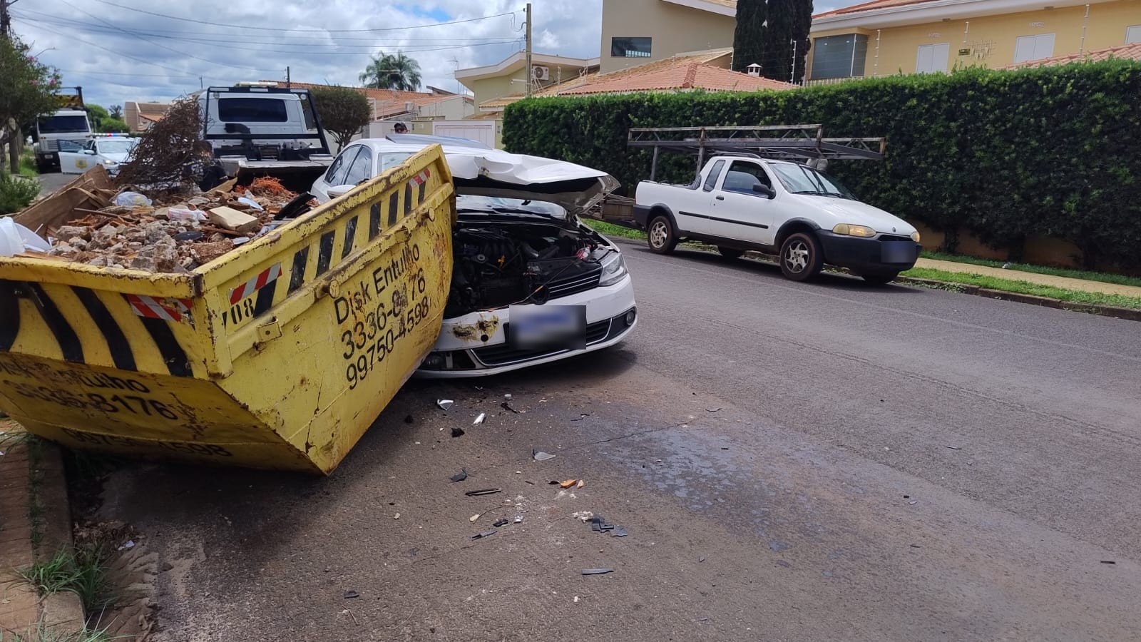Sedã branco com a frente destruída encostado em uma caçamba amarela de entulho, com peças espalhadas no asfalto