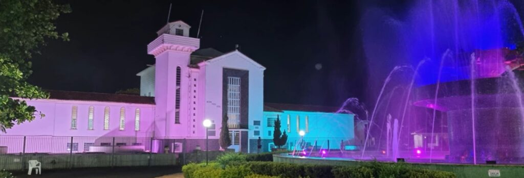 Prédio branco da sede do Daae iluminado à noite em tons de rosa e azul, com um chafariz ao lado