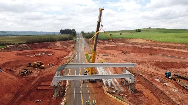 Vista aérea de guindaste erguendo uma viga de concreto sobre a pista da SP-255, com terra vermelha da obra dos dois lados