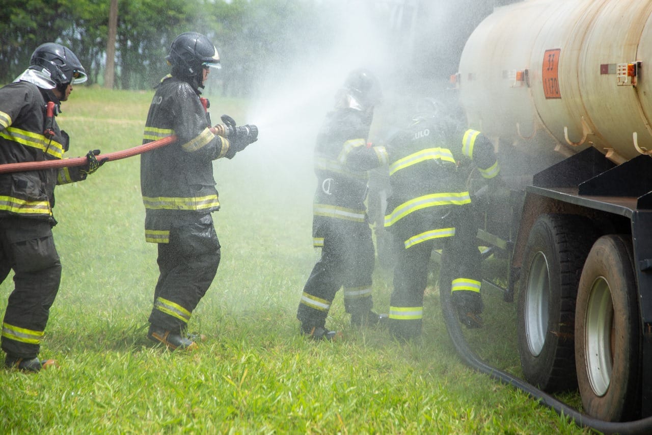 Bombeiros com roupa de proteção jogam jatos de água sobre um caminhão-tanque parado no gramado
