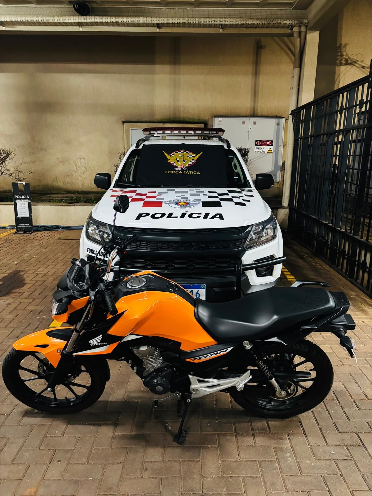 Policiais da Força Tática ao lado de uma moto laranja estacionada na garagem de uma residência.